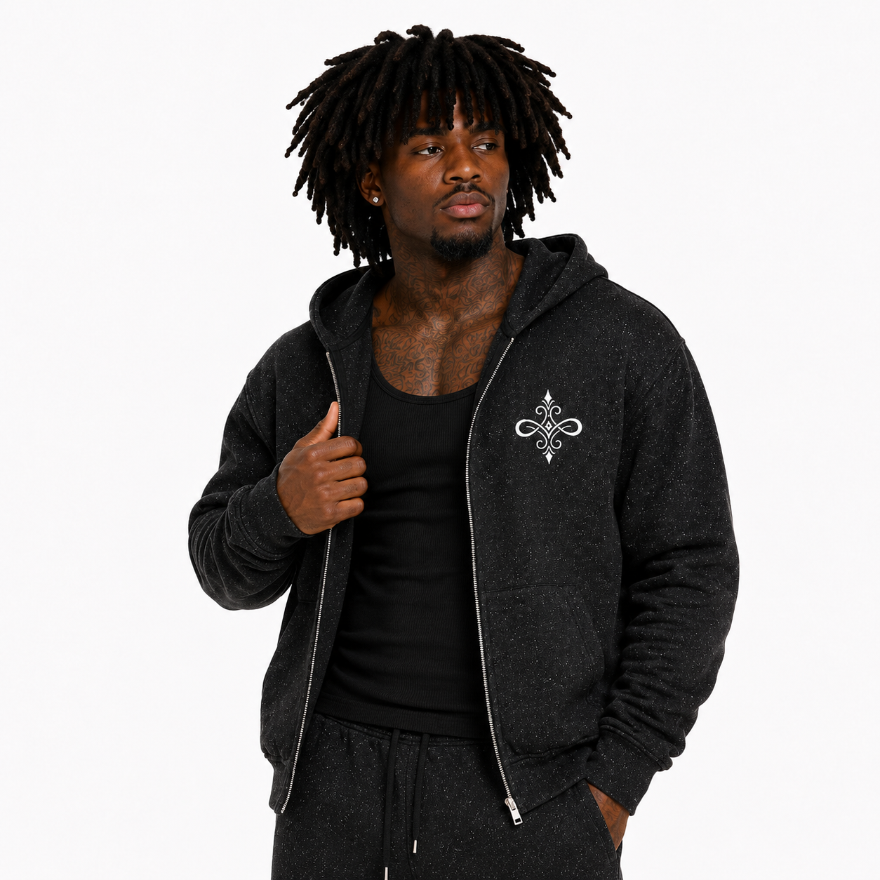 A man with dreadlocks wearing a dark charcoal gray zip-up hoodie with a white ornamental emblem on the left chest, styled over a black tank top and matching dark sweatpants, set against a plain white background.