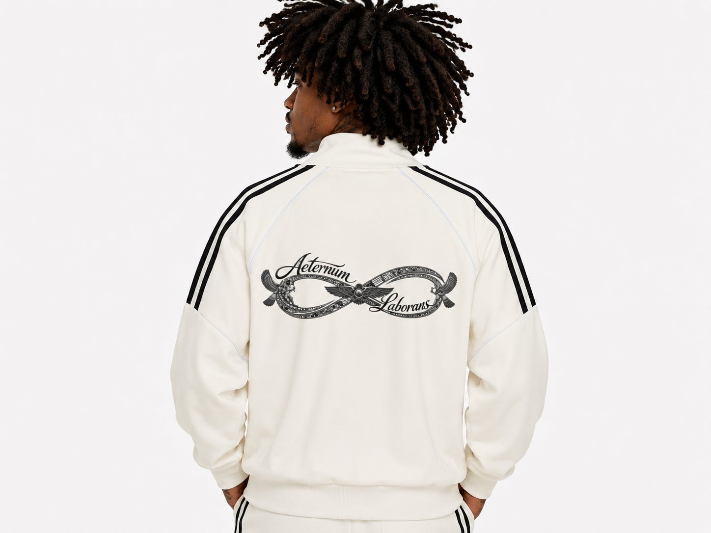 A man viewed from behind wearing a cream-colored track jacket with black stripes on the shoulders and a large black graphic on the back, set against a plain white background.