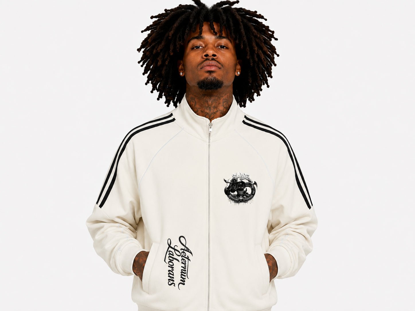 A man with dark curly hair wearing a cream-colored zip-up track jacket with black shoulder stripes and black graphic prints on the chest and sleeve, set against a plain light gray background.