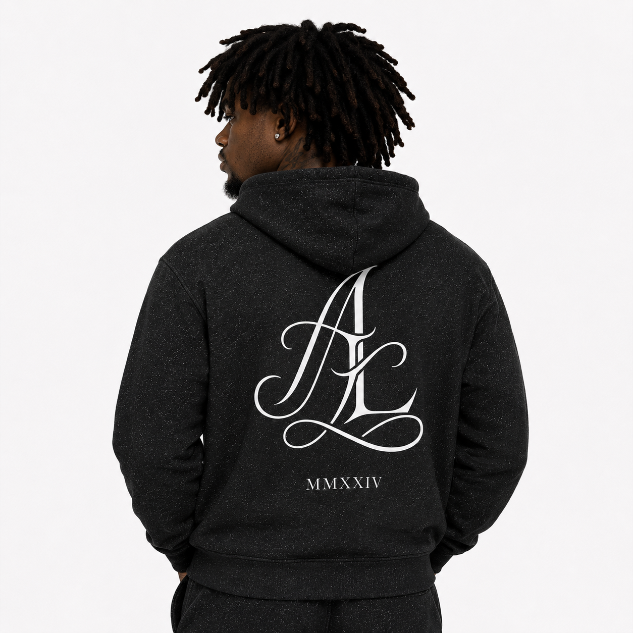 A man viewed from the back wearing a dark charcoal gray hoodie with a large white cursive 'AK' monogram and 'MMXXIV' text printed on the back, set against a plain light gray studio background.