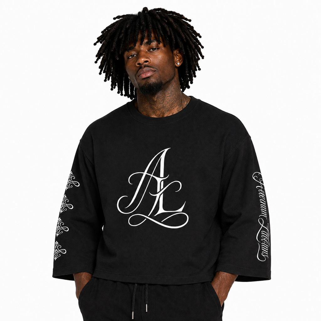 A man with black dreadlocks wearing a black oversized crewneck sweatshirt with a large white 'AK' monogram on the chest and white script graphics on the sleeves, paired with black sweatpants against a plain white background.