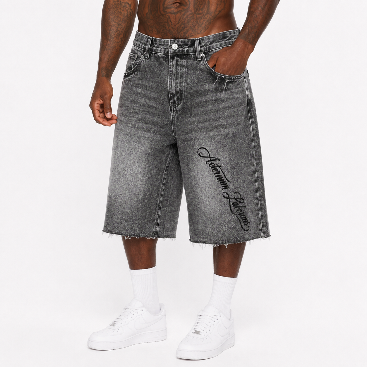 A model wearing gray acid-wash denim shorts with a black script logo on the left leg, styled with white crew socks and white sneakers against a plain white background.