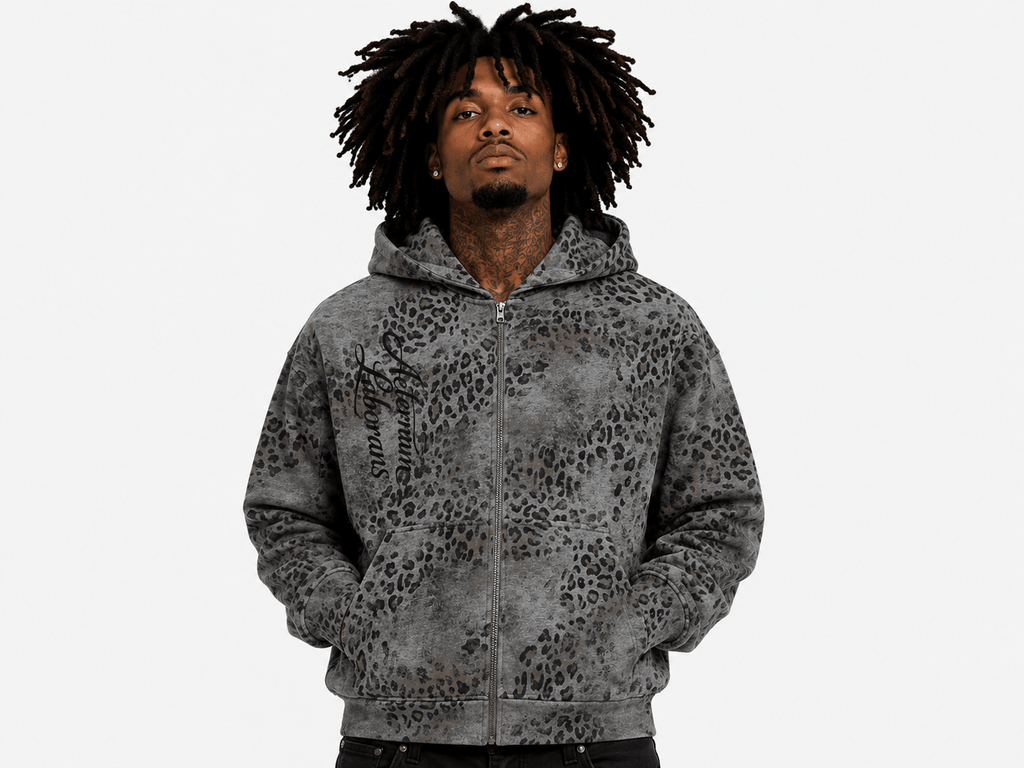 A man with dark dreadlocks wearing a gray and black leopard-print zip-up hoodie, photographed against a solid light gray background.