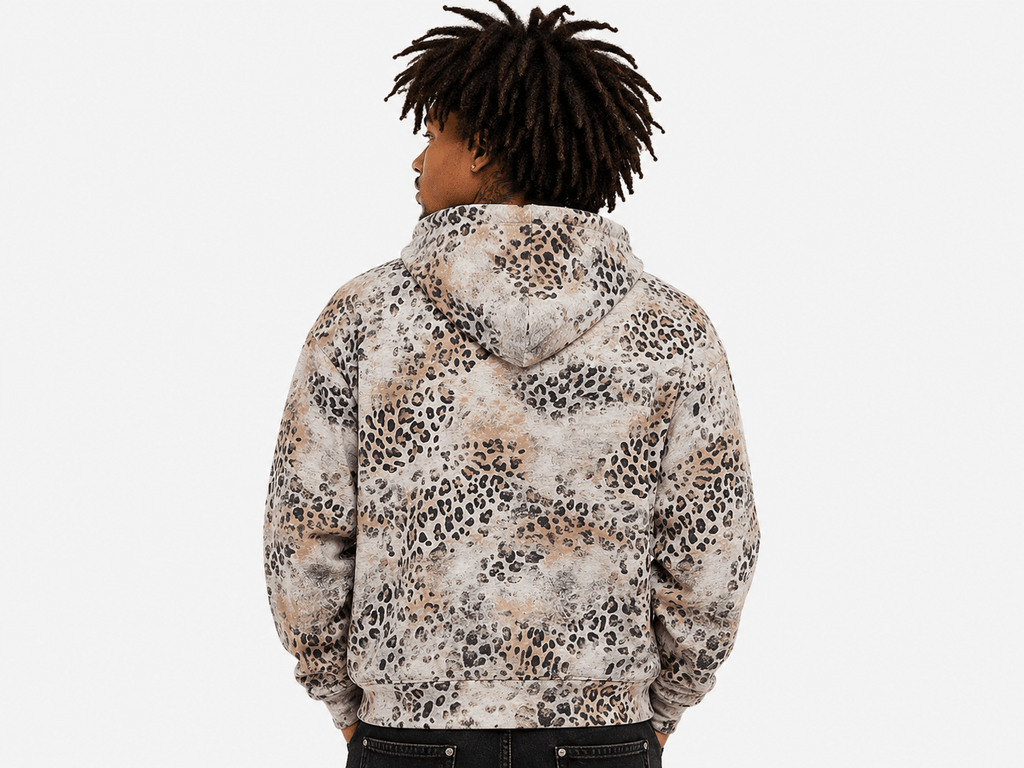 A person shown from the back wearing a hooded sweatshirt with an allover leopard print pattern in beige, brown, black, and off-white, against a plain light gray background.