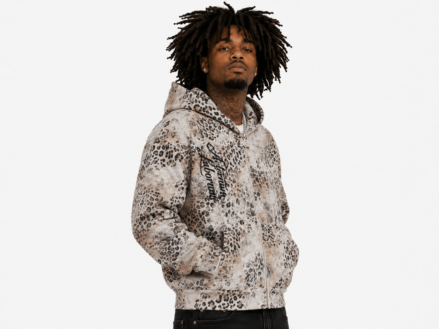 A man with dark curly hair wearing a beige and brown leopard print zip-up hoodie, standing against a solid white background.