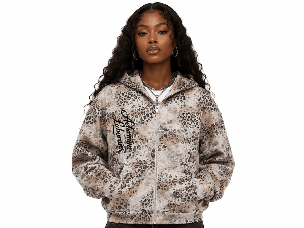 A woman modeling a zip-up hoodie with an animal print pattern in beige, brown, and black, featuring a black script graphic on the left chest, set against a plain white background.