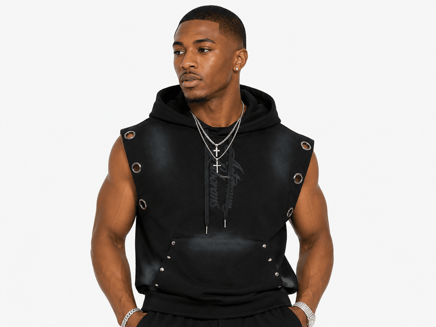 A man wearing a black sleeveless hooded vest with metal grommets and drawstrings, styled with silver chain necklaces, against a plain light gray background.