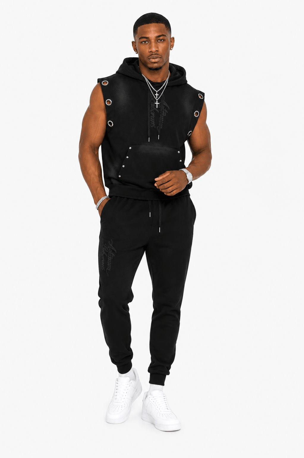 A man modeling a black sleeveless zip-front vest with faux-leather panels, metal grommets, and a hood, paired with black jogger pants and white sneakers, against a light gray studio background.