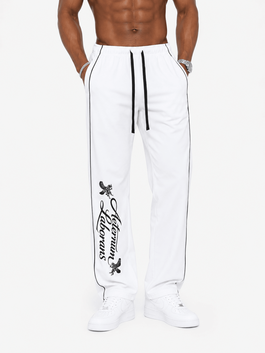 White jogger sweatpants with black side piping, a black drawstring, and a black vertical script logo on the left leg, shown on a model against a light gray background.