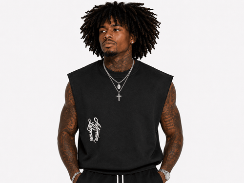 A man with dark curly hair wearing a black sleeveless t-shirt with a white graphic on the left chest, styled with silver necklaces, against a light gray background.