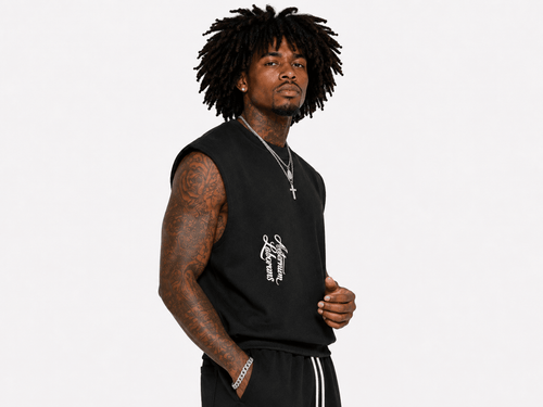A man with dark curly hair wearing a black sleeveless t-shirt with a white graphic on the chest, paired with black track pants, photographed against a solid light gray background.