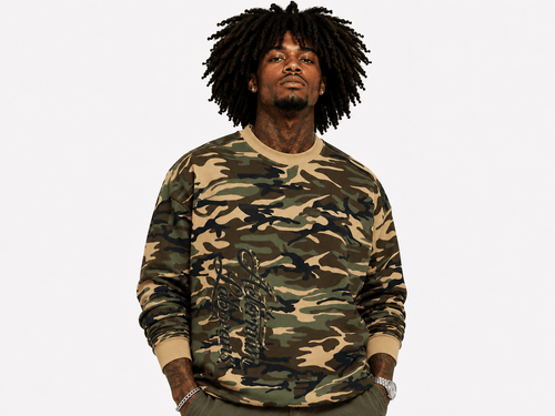 A man with dark dreadlocks wearing a camouflage crewneck sweatshirt with beige ribbed collar, cuffs, and hem, posed against a solid white background.