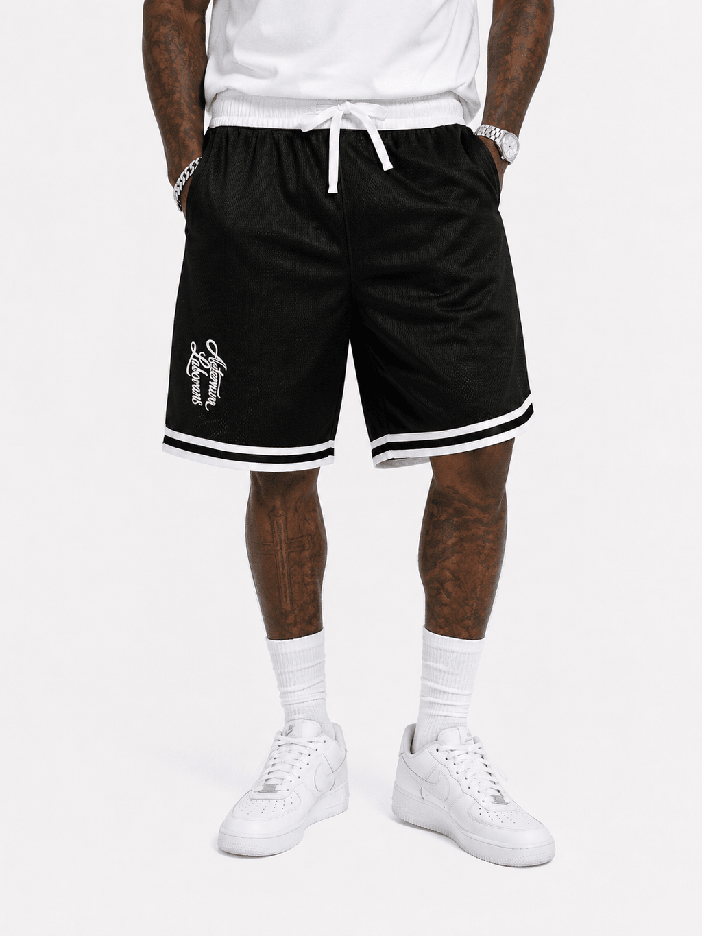 Black basketball shorts with white trim and a white drawstring, featuring a small white graphic on the left leg, modeled with a white t-shirt, white socks, and white sneakers.