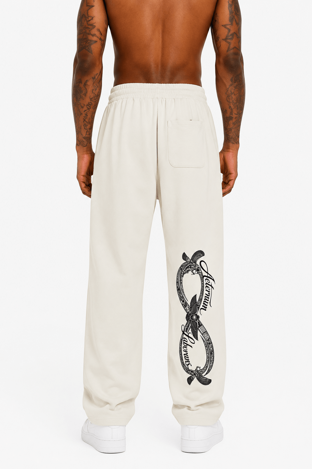 A man shown from the back wearing cream-colored sweatpants with a black graphic print on the left leg, paired with white sneakers against a light gray studio background.