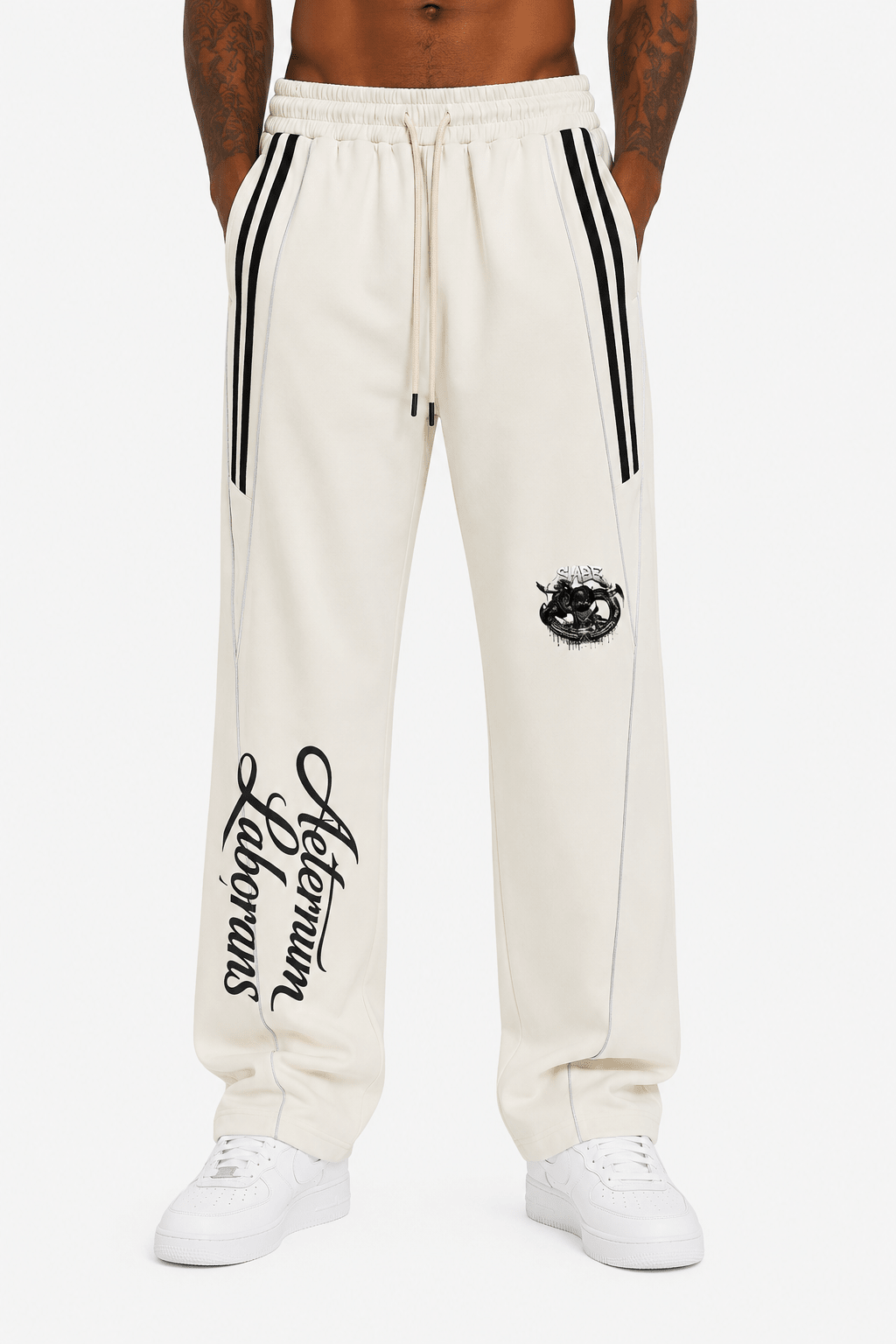 A person wearing off-white sweatpants with black side stripes, a black embroidered crest on the thigh, and black script text down one leg, paired with white sneakers against a light gray studio background.
