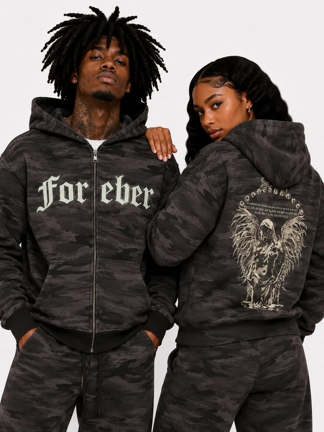 Two people wearing matching camouflage hoodies with text and graphics on a plain background