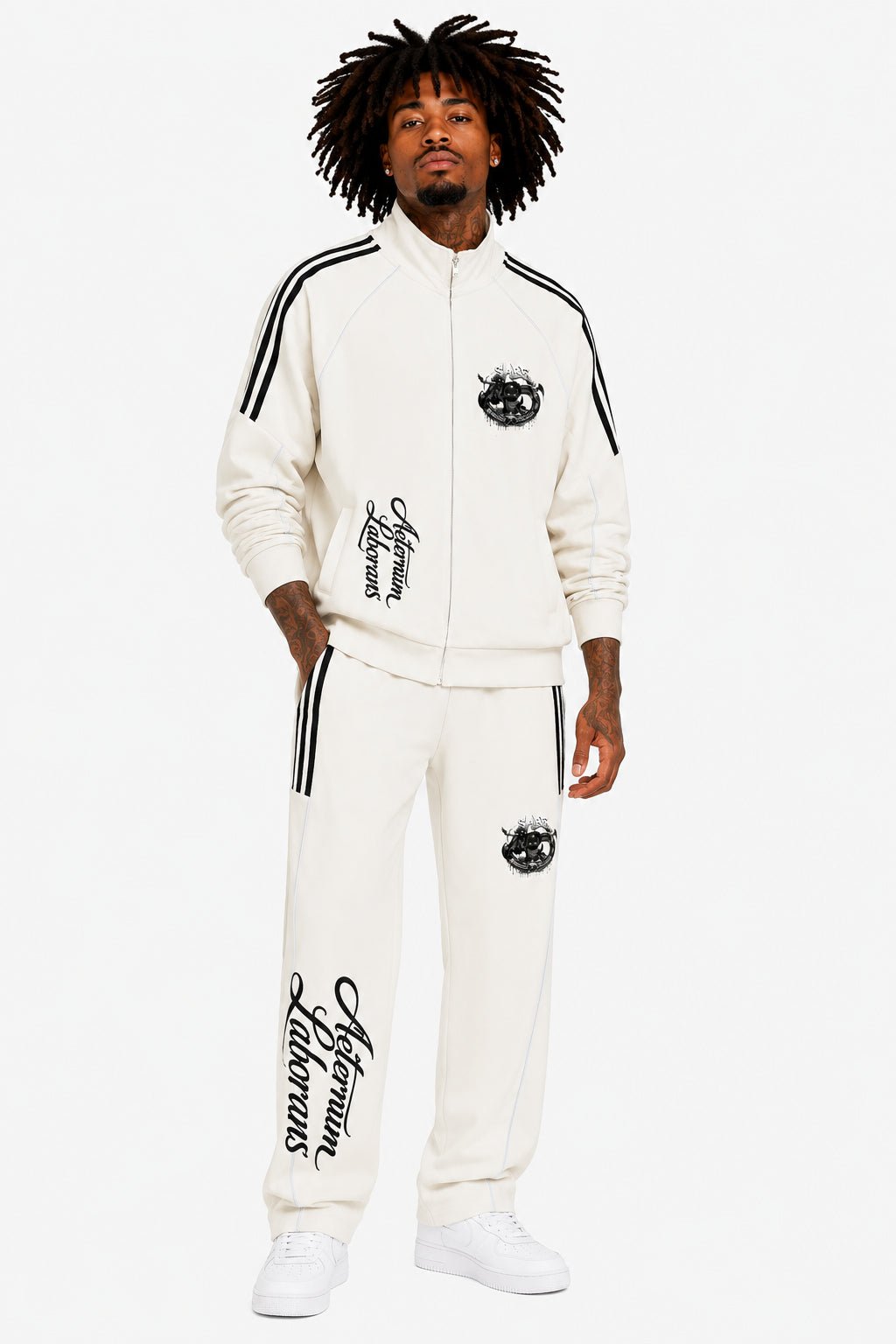 A man modeling a matching off-white tracksuit with black graphics, including a zip-up jacket with shoulder stripes and sweatpants with script text and a crest logo, against a plain white background.