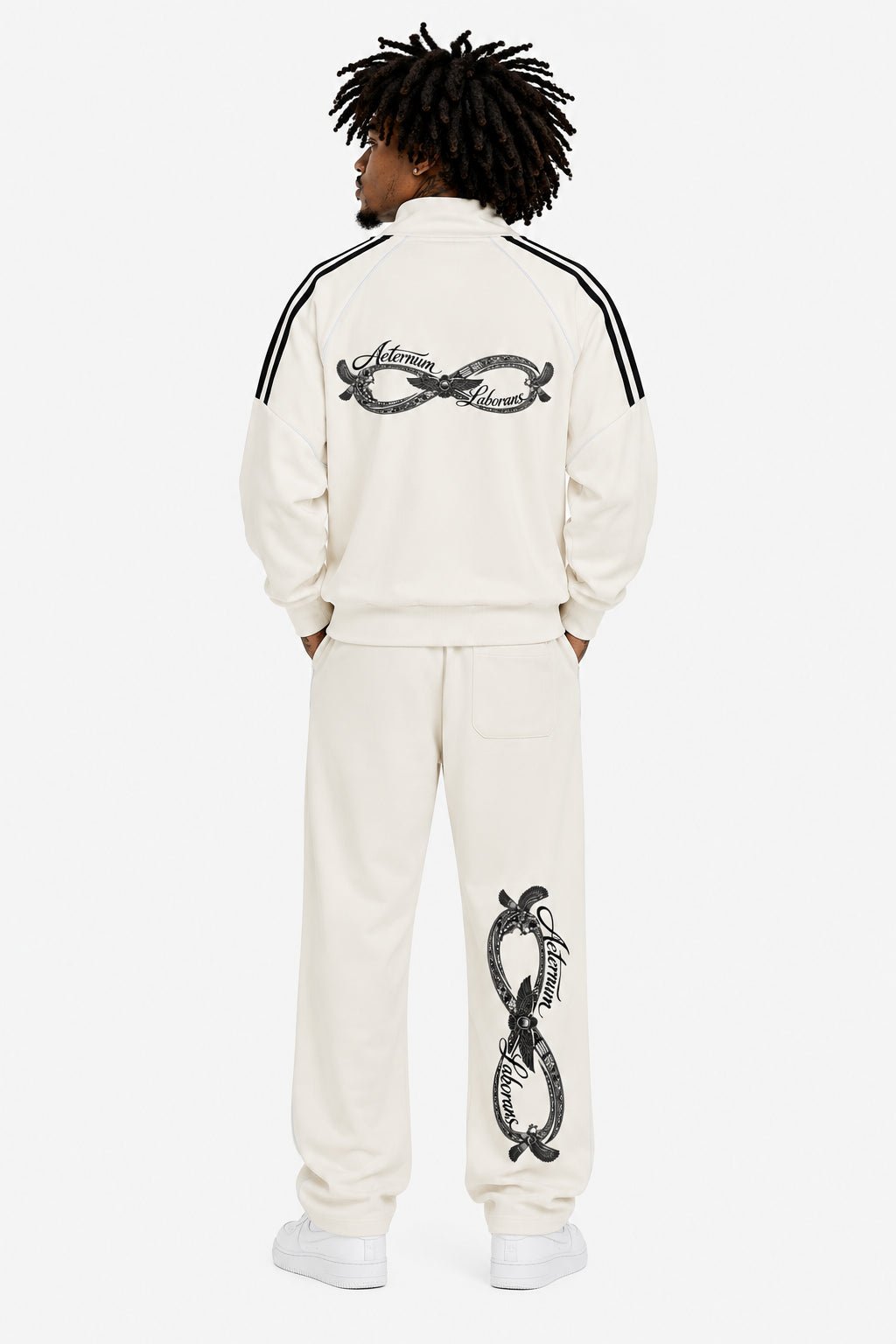 A model shown from the back wearing a cream-colored tracksuit with black details. The jacket features a black infinity symbol printed across the upper back, and the matching sweatpants have a large black snake graphic wrapped around an infinity symbol on the back of the leg.